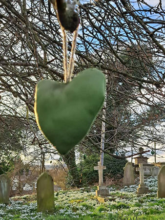 A felt green heart hangs from a tree in a churchyard with snowdrops on the ground