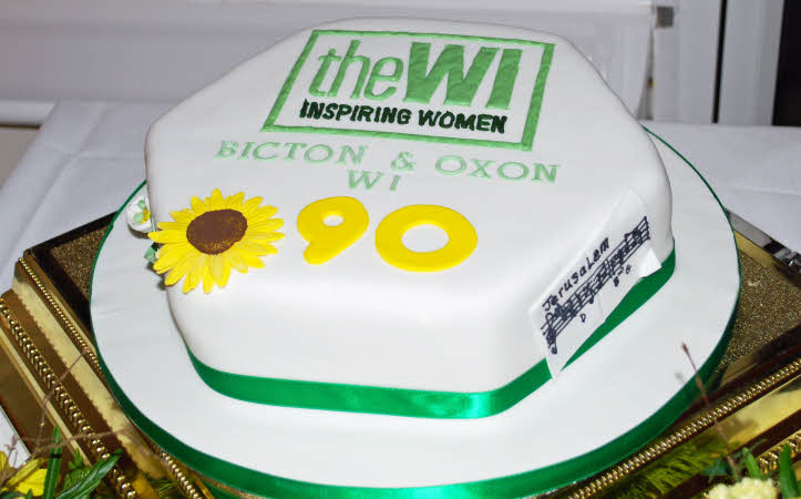 A hexagonal cake, with white icing, a green ribbon, the WI logo, our branch name, 90 in yellow and a daisy. The first few bars of Jerusalem are pinned to the side.