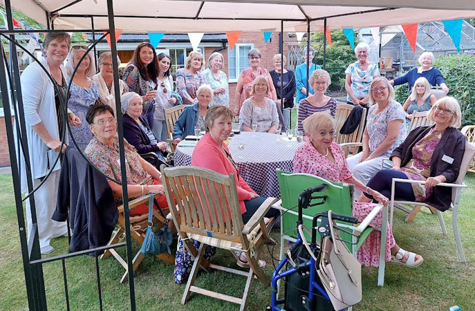 Twenty two ladies stand or sit around a table under a gazebo, all face the camera